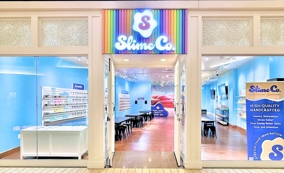 Slime Co. retail store entrance in a shopping mall, featuring a colorful rainbow sign with a white slime blob logo, bright blue walls, display shelves, and tiled flooring.